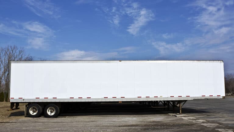 Side view of a long, white, unbranded semi-trailer parked in a lot under a blue sky.