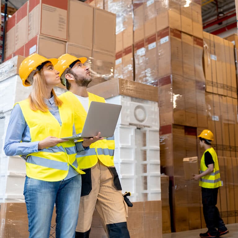 Two warehouse managers wearing yellow hard hats and high-visibility safety vests standing in a large distribution center.