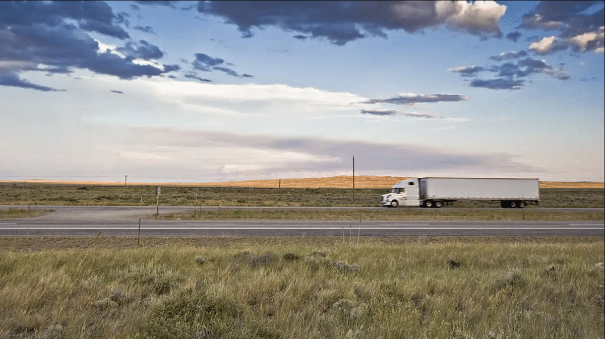 White semi-truck and trailer driving along a rural highway with open grassy fields and a blue sky background.