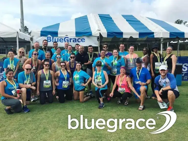 A large group of smiling BlueGrace Logistics employees wearing blue and grey athletic gear and medals, posing under a blue and white striped tent after a community race.