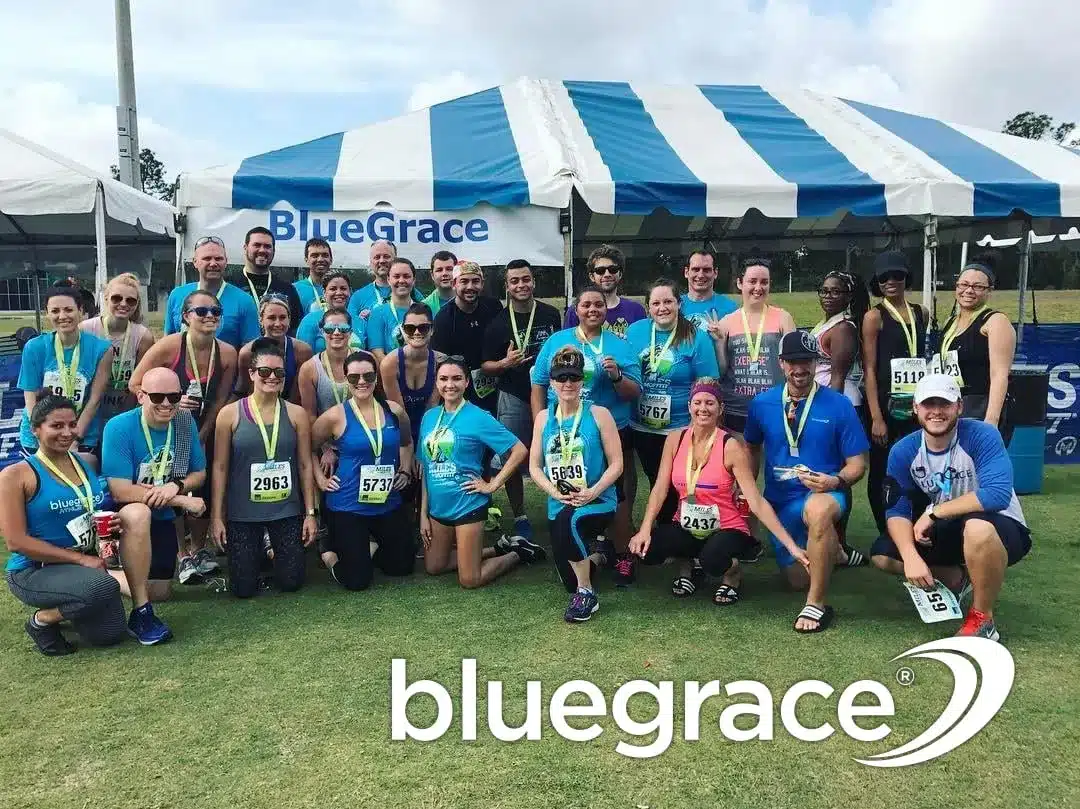 A large group of smiling BlueGrace Logistics employees wearing blue and grey athletic gear and medals, posing under a blue and white striped tent after a community race.