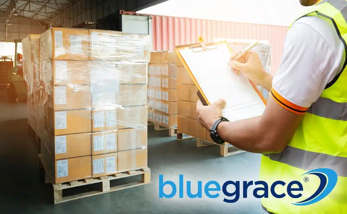 Warehouse worker in a yellow safety vest writing on a clipboard next to stacks of plastic-wrapped cardboard boxes.