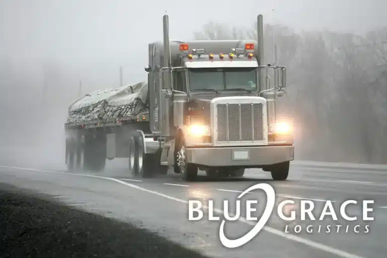 Front view of a semi-truck driving on a highway through thick fog with headlights illuminated.