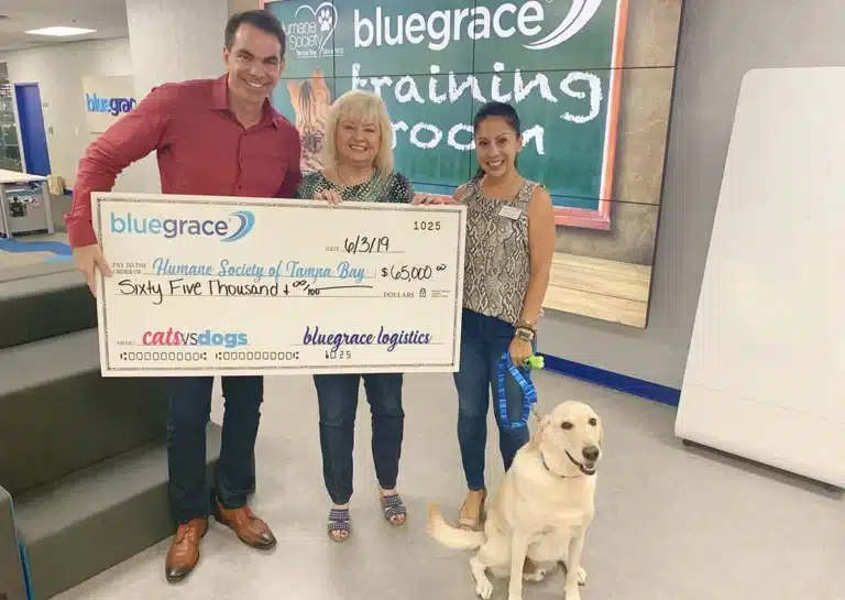 Three smiling professionals from BlueGrace Logistics presenting a large ceremonial check for $65,000 to the Humane Society of Tampa Bay, accompanied by a yellow Labrador retriever.