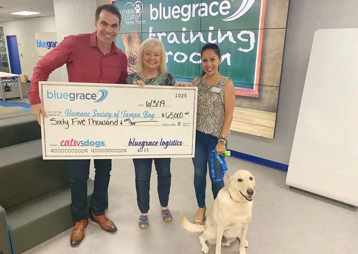 Three smiling professionals from BlueGrace Logistics presenting a large ceremonial check for $65,000 to the Humane Society of Tampa Bay, accompanied by a yellow Labrador retriever.