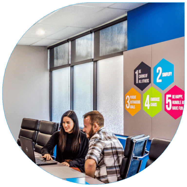 A professional workspace featuring two employees collaborating on a laptop in a modern conference room, adjacent to a wall display of the company's five core values.