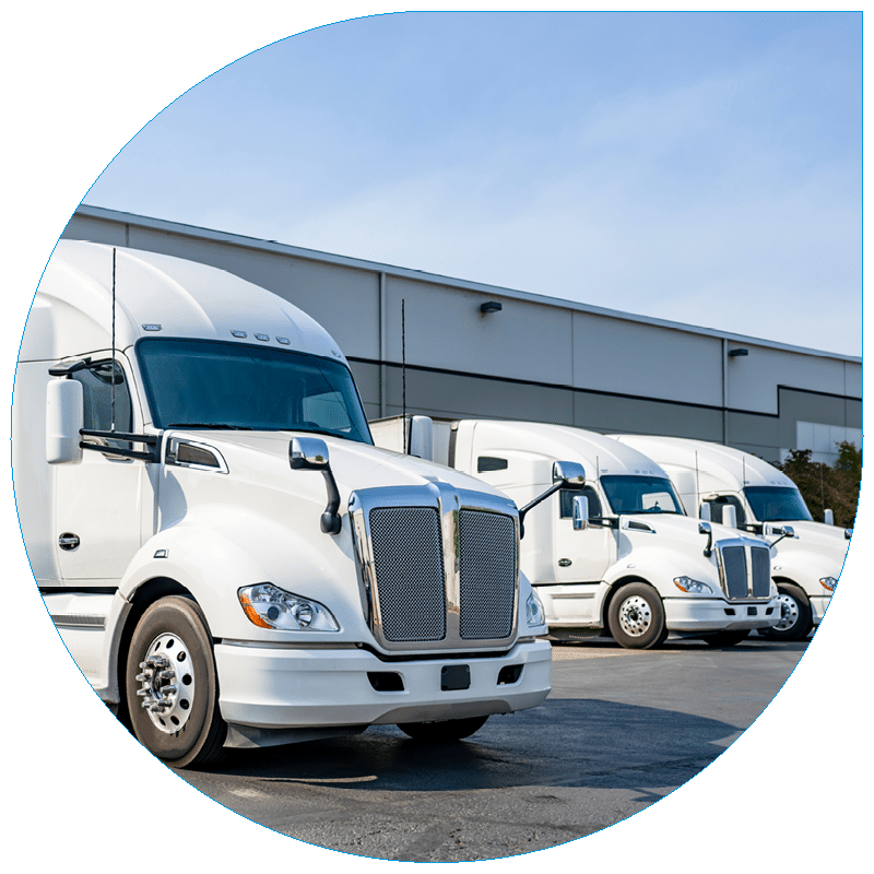 Three modern white semi-trucks parked in a row outside a large industrial warehouse facility during the day.