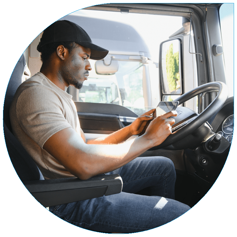 A truck driver in a tan t-shirt and black baseball cap sitting in the cab of a modern semi-truck, focused on using a digital tablet for route or shipment management.