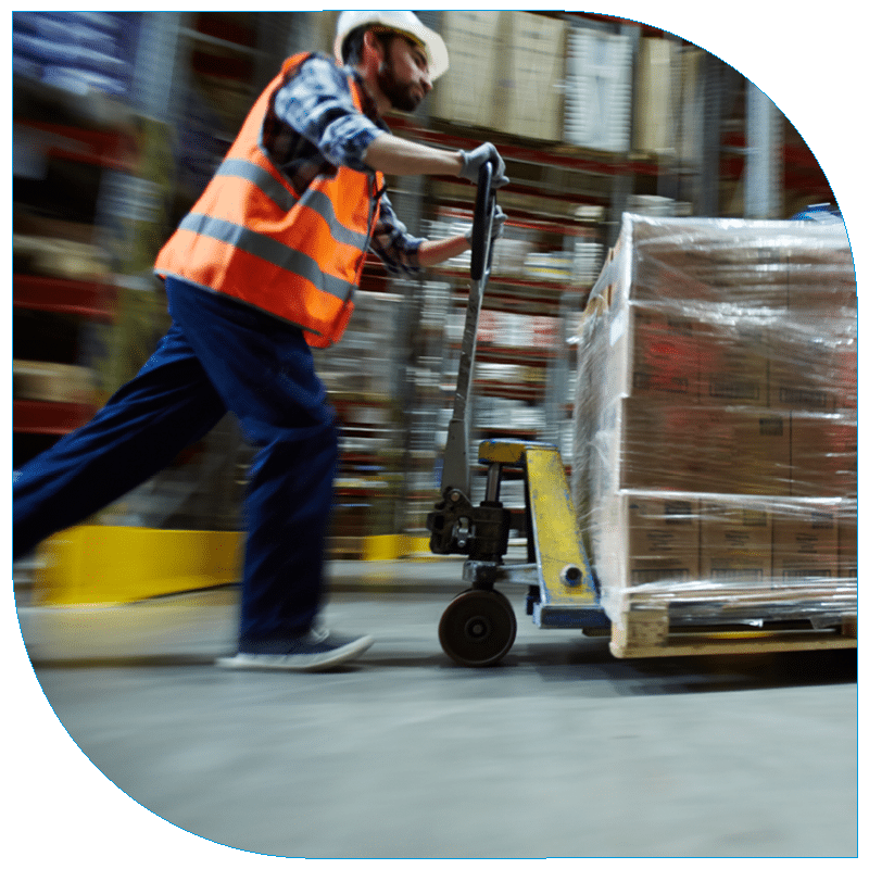 Motion-blur side view of a warehouse worker in an orange safety vest and white hard hat quickly pushing a manual pallet jack with shrink-wrapped boxes through a storage facility.