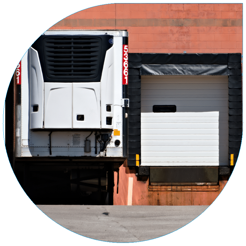 Exterior view of a white refrigerated semi-trailer, or reefer, backed into a warehouse loading dock next to a closed bay door.