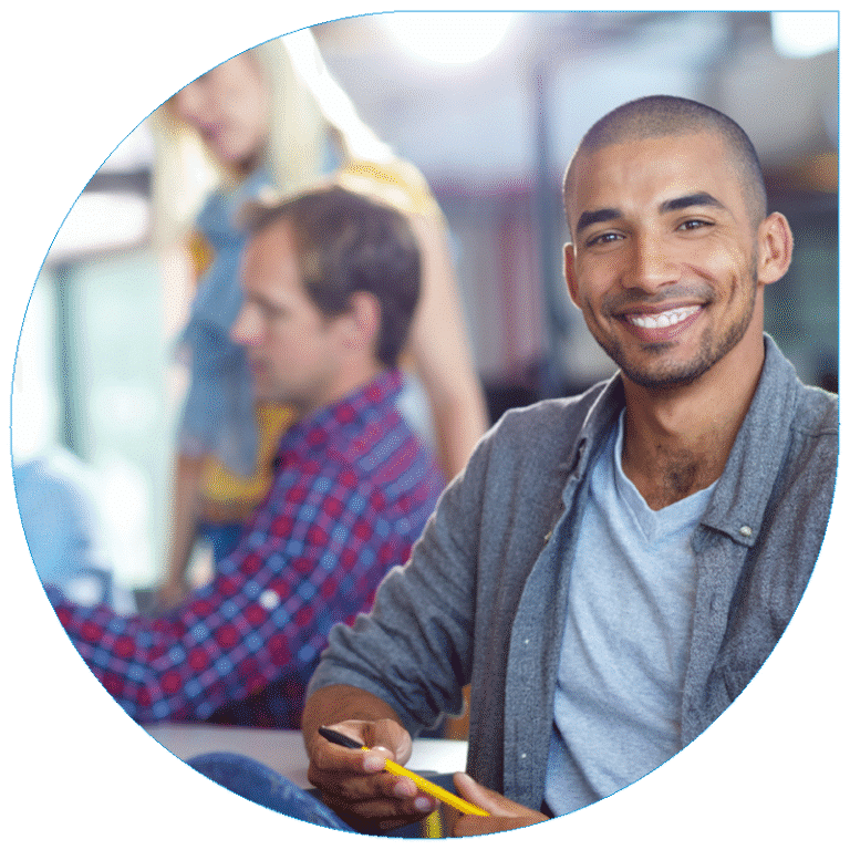 Portrait of a smiling young professional man sitting at a desk in a busy creative office.