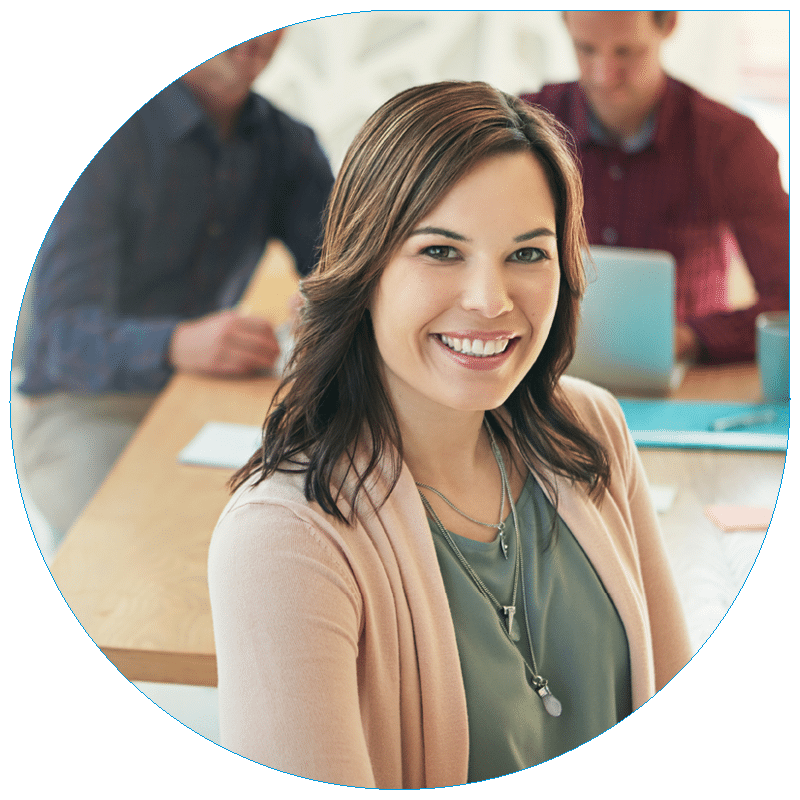 Portrait of a smiling professional woman wearing a pink cardigan sitting at a conference table with colleagues in the background.