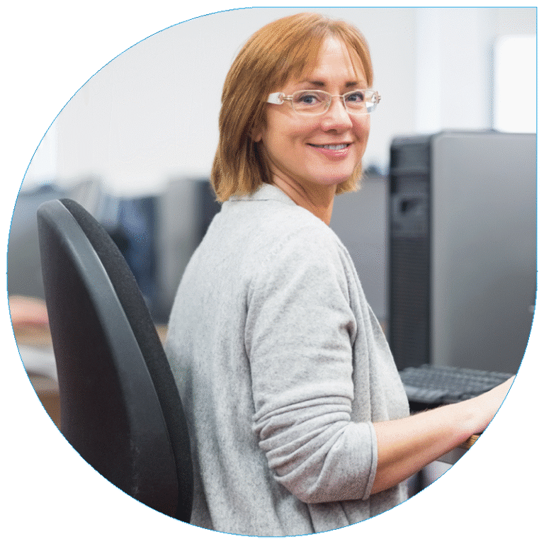 Portrait of a smiling professional woman with glasses and short reddish-brown hair sitting at a computer desk in an office.