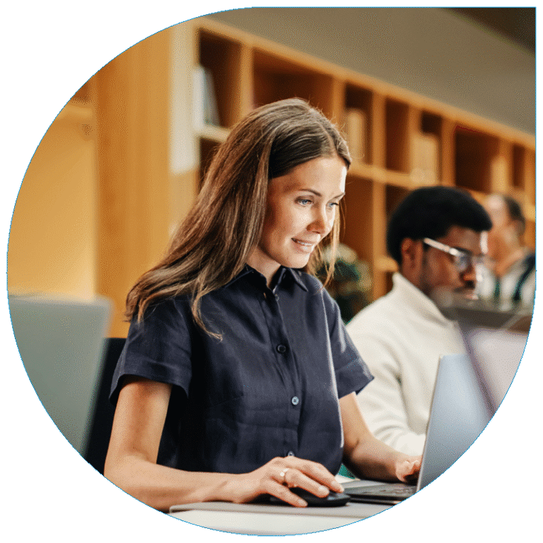 Portrait of a smiling professional woman with long brown hair using a laptop and mouse in a modern, open-concept office.