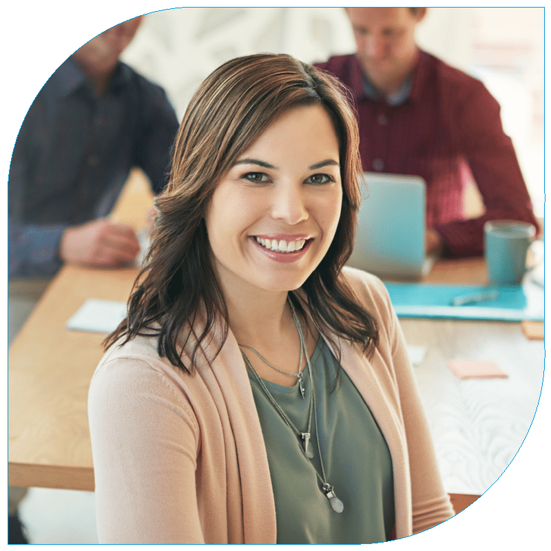 Portrait of a smiling professional woman wearing a pink cardigan sitting at a conference table with colleagues in the background.
