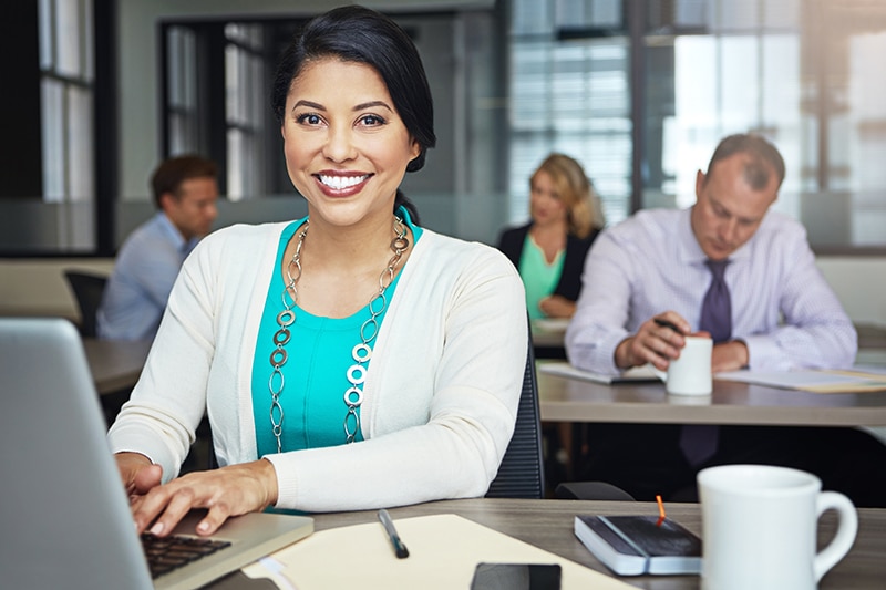 Smiling woman in a teal top and white cardigan using a laptop in a modern professional office.