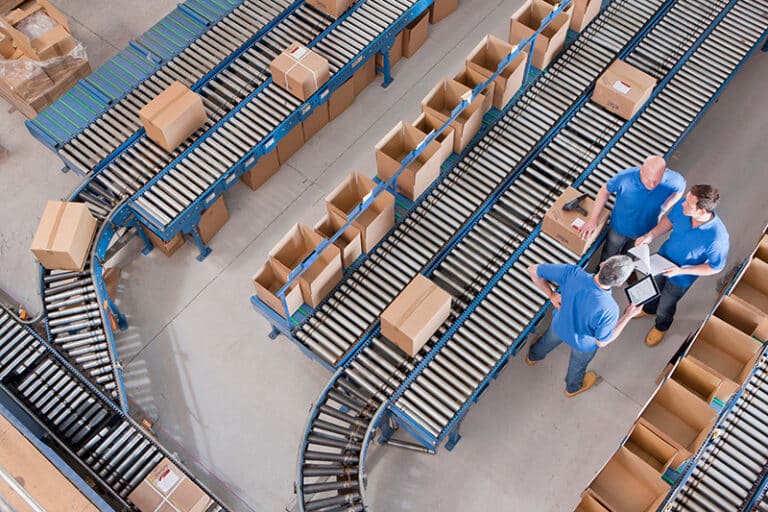 Top view of workers with papers and a digital tablet having a discussion among boxes laid on conveyor belts at a distribution warehouse
