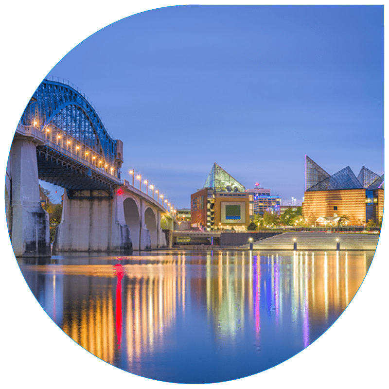 Illuminated blue metal bridge spanning a Chattanooga river at dusk, with modern architectural buildings reflecting in the water.