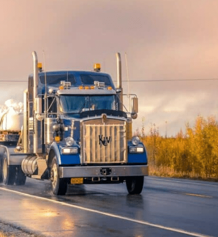 A large blue semi-truck with chrome accents and amber roof lights traveling on a wet asphalt road at sunset, with a line of trees in the background.