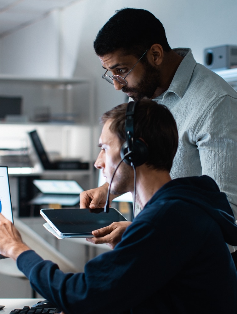 A senior analyst with glasses standing over a colleague wearing a headset.