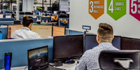 Employee working at a multi-monitor desk setup in an open office, next to a wall displaying colorful company core values graphics.